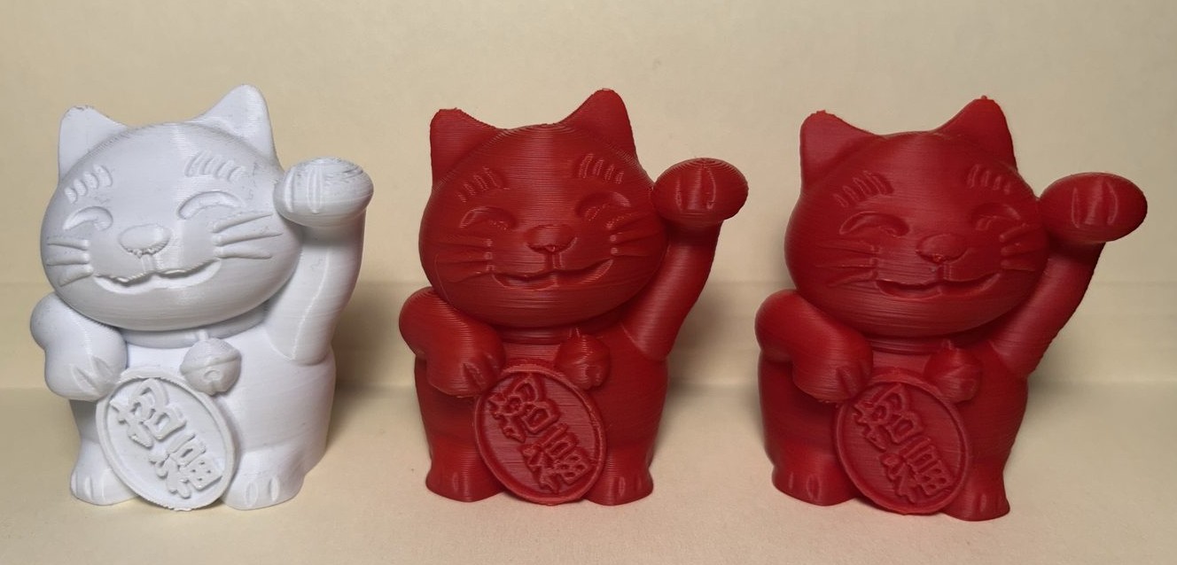 Three 3D-printed lucky cat models side by side: the white original from the stock machine, and two red prints from the rebuilt one